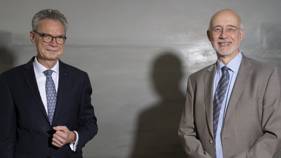 Herr Lutz steht links und trägt eine Brille und einen dunklen Anzug. Herr Puhl steht rechts und trägt eine Brille und einen grauen Anzug