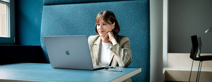 Frau mit braunen Haaren und Blazer sitzt mit Laptop in einer türkisfarbenen Arbeitsecke.