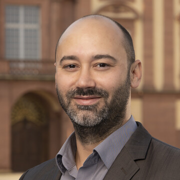 Jochen Baumgardt is bald, has a black and gray beard, wears a gray shirt and a brown jacket and is standing in front of the castle.