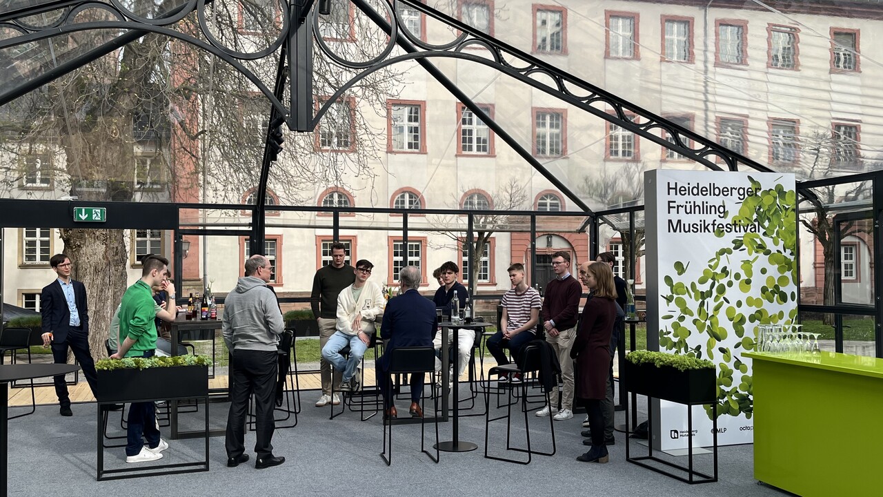 Personen, Stehtische und Stühle unter einem gläsernen Pavillon. Auf einem Poster steht: Heidelberger Frühling Musikfestival