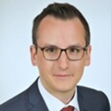 Herr Strohm hat kurze schwarze Haare und trägt eine weißes Hemd, blaues Sakko und eine rote Krawatte