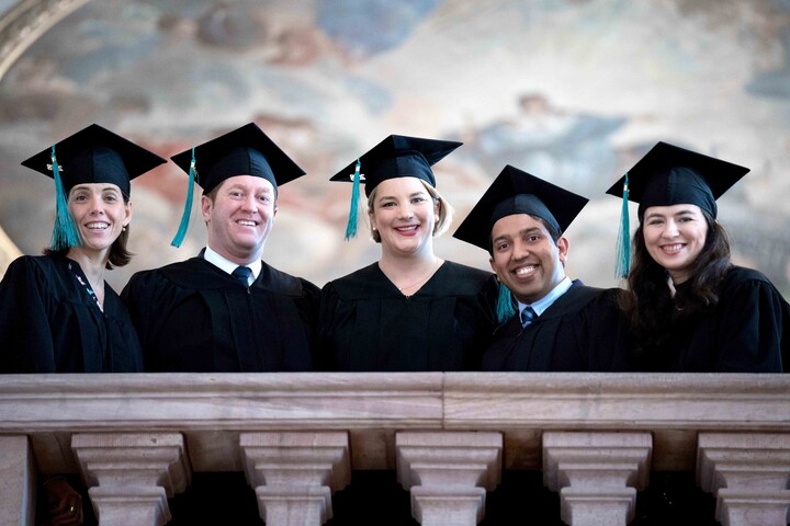 Five people stand next to each other on a balcony. They are wearing gowns and hats.