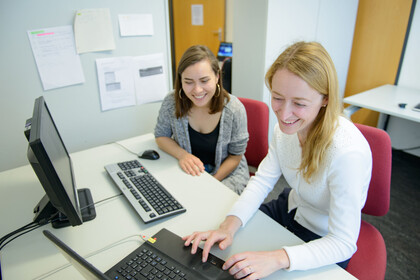 Zwei Frauen sitzen lachend an einem Tisch nebeneinander vor einem Laptop und einem Bildschirm mit einer externen Tastatur.