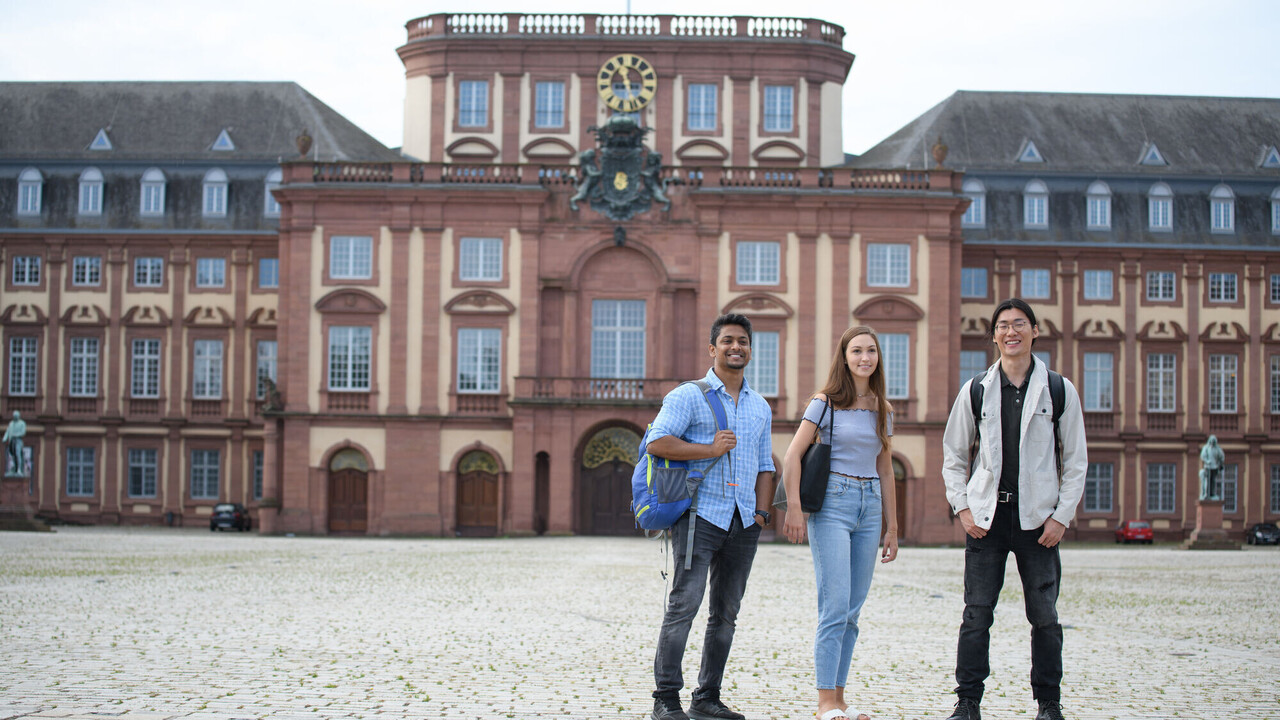 Students at the Ehrenhof Three students in the castle courtyard look towards the camera. The central building of the palace in the background.