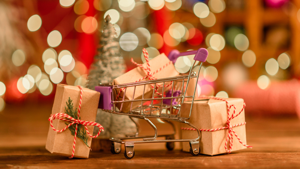 Ein Mini-Einkaufswagen gefüllt mit Geschenken, umgeben von weiteren Geschenkpaketen, vor einem verschwommenen, festlich beleuchteten Hintergrund.