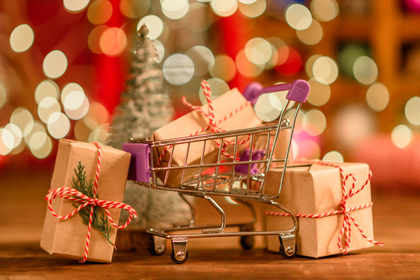 Ein Mini-Einkaufswagen gefüllt mit Geschenken, umgeben von weiteren Geschenkpaketen, vor einem verschwommenen, festlich beleuchteten Hintergrund.