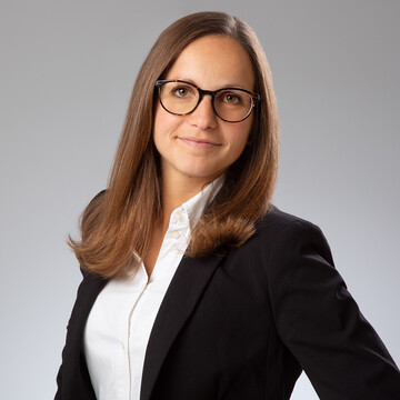 Annabelle Bröstl. Sie trägt ein schwarzes Kostüm und steht vor einer grauen Wand.