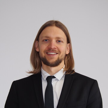 Herr Kinski hat schulterlange braune Haare und trägt ein weißes Hemd mit schwarzem Blazer und Krawatte
