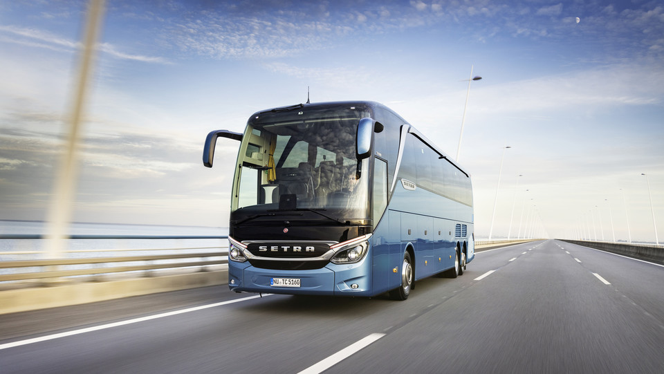 Ein blauer Reisebus fährt über eine Brücke. Im Hintergrund das Meer.