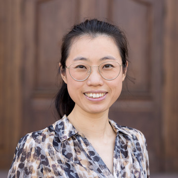 Yufang Sun. Sie trägt eine geblümte Bluse und steht vor einer Holztür.