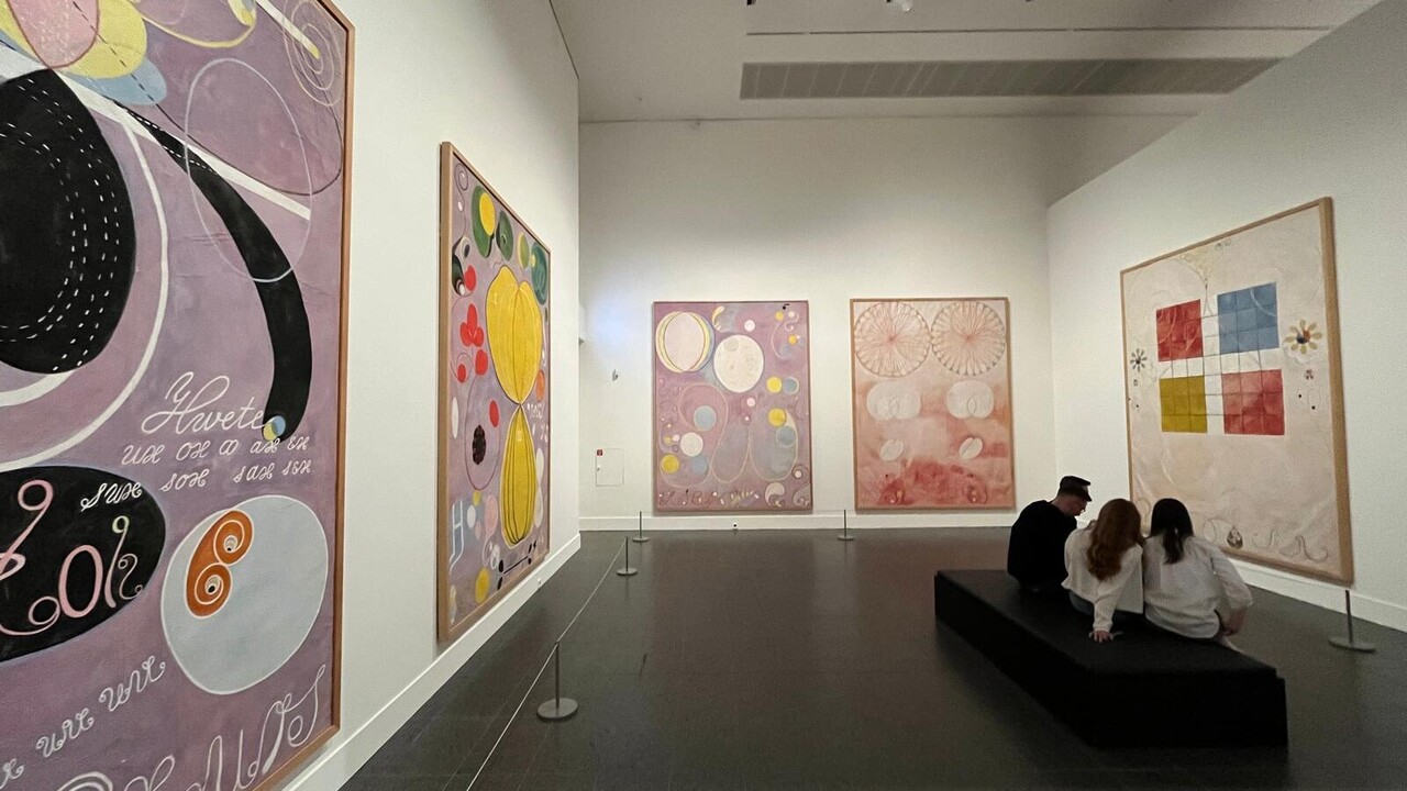 Raum einer Kunstgalerie. An den Wänden hängen Bilder, in der Mitte ist eine Wand auf der Personen sitzen