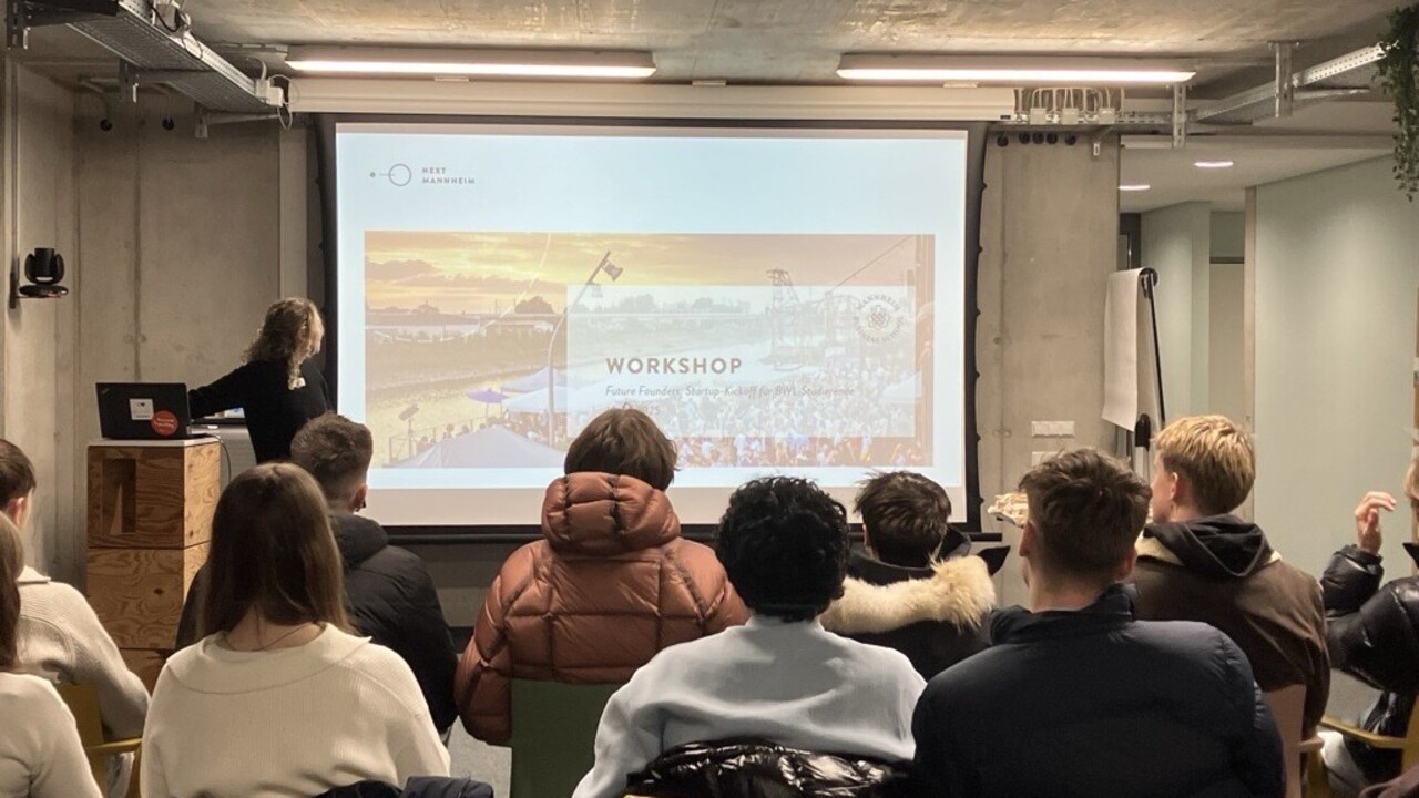 Eine Personengruppe von hinten. Die Menschen sitzen auf Stühlen und schauen auf eine Präsentation.