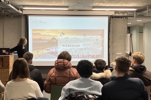 Eine Personengruppe von hinten. Die Menschen sitzen auf Stühlen und schauen auf eine Präsentation.