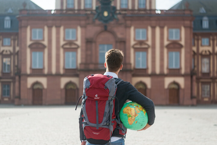 Person mit Rucksack und Globus unterm Arm vor dem Schloss von hinten