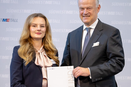 Frau Zercher und Herr Krause stehen vor einer Wand. Sie halten zusammen eine Urkunde und lächeln in die Kamera.
