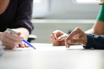 Internationale Studierende Zwei Studierende werden beraten, dabei sind die Hände und der Stift der Mitarbeiterin, der auf ein Papier zeigt, im Fokus.