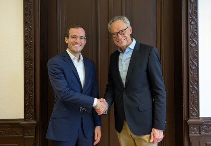 Dekan Lutz und Prof. Reinwald stehen vor einer dunklen Holztür und schütteln sich die Hände. Beide Lächeln in die Kamera.