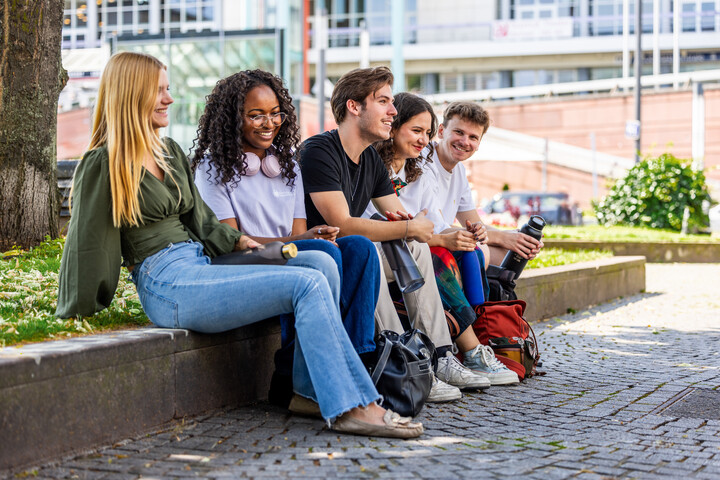 Fünf Studierende sitzen auf einer Bank im Park. Sie lachen gemeinsam.