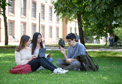 Drei Studierende sitzen auf dem Campus auf der Wiese. Sie haben Unterlagen und Laptops vor sich aufgeschlagen.