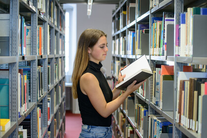 Eine Studentin steht zwischen zwei Bücherregalen und liest in einem Buch. 