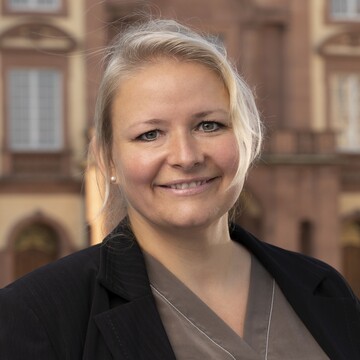 Ines Beese hat blonde Haare in einem Zopf. Sie trägt eine beige Bluse und einen dunklen Blazer. Im Hintergrund ist das Schloss zu sehen.