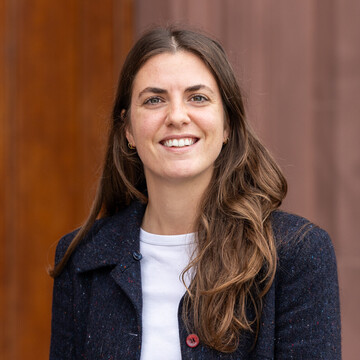 Linda hat langes braunes Haar und trägt ein weißes T-Shirt mit einer dunkelblauen Jacke. Im Hintergrund ist das Schloss zu sehen.