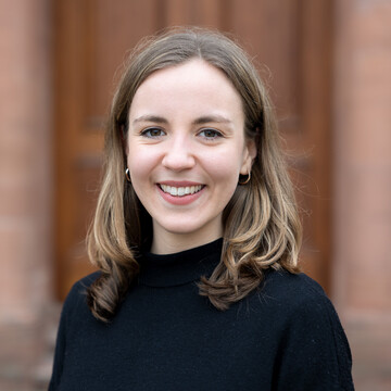 Lena Rudat. Sie trägt einen schwarzen Pullover und steht vor einer Holztür.