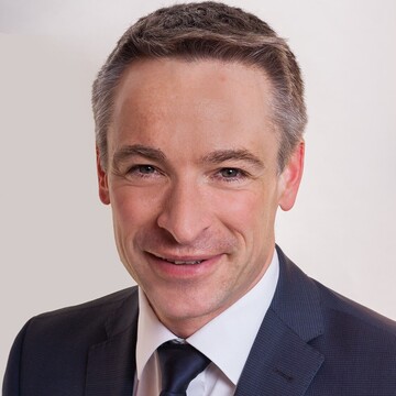 Herr Schlipf hat braune kurze Haare und trägt ein weißes Hemd mit blauem Anzug und blauer Krawatte