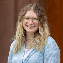 Porträt von Franziska Jäger. Franziska Jäger trägt ein hellblaues Hemd und eine beige Brille mit großen Gläsern. Ihre dunkelblonden Haare fallen offen über die Schultern. Sie lacht herzlich und trägt mehrere goldfarbene Halsketten.