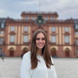 Michelle Back has long brown hair and is wearing a white blouse. The palace and Ehrenhof can be seen in the background.