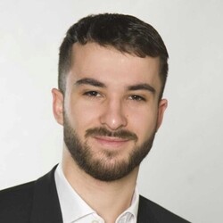 Tim Jonas has short dark hair and a beard. He is wearing a dark jacket and a white shirt.