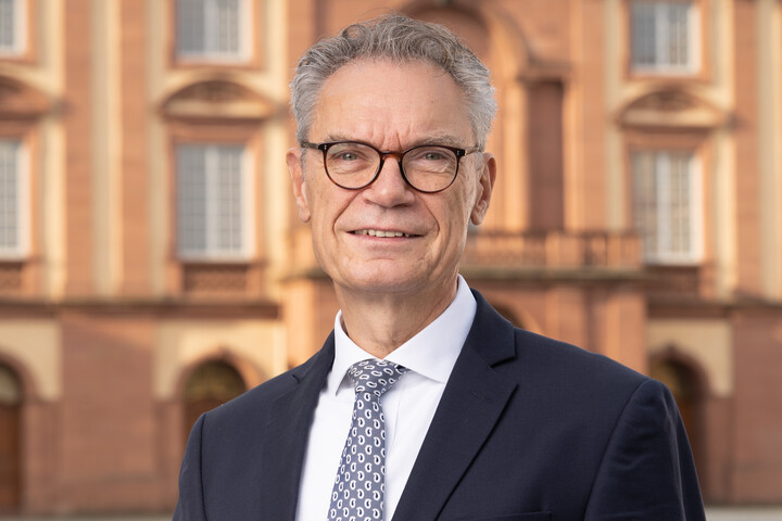 Joachim Lutz trägt einen Anzug und hat graue Haare und eine Brille. Hinter ihm ist das Schloss zu sehen
