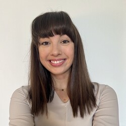 Hannah Bauer has long dark hair and bangs. She wears a white top and smiles at the camera.