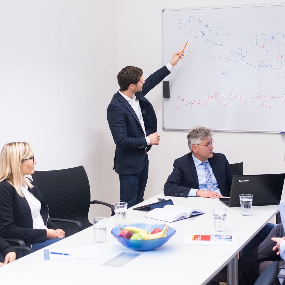 Personengruppe sitzt um Tisch, jemand erklärt Darstellungen an Whiteboard