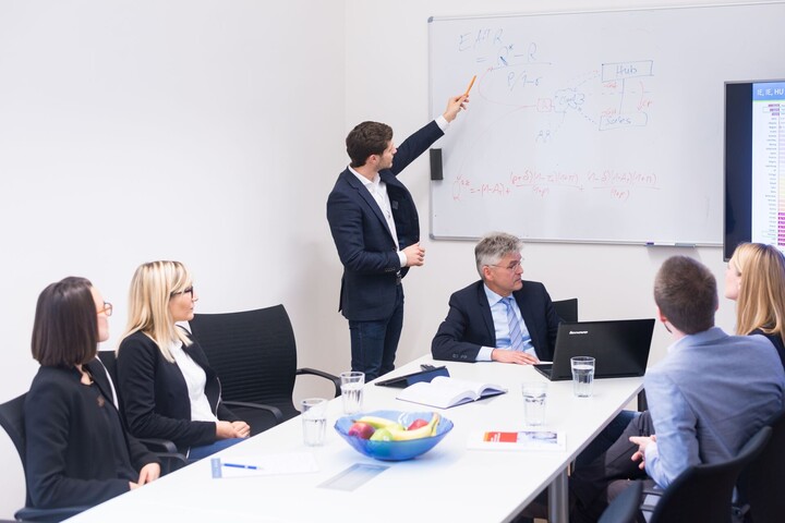 Personengruppe sitzt um Tisch, jemand erklärt Darstellungen an Whiteboard